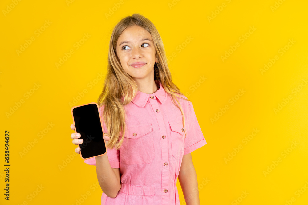 Beautiful caucasian kid girl wearing pink shirt holds new mobile phone and looks mysterious aside shows blank display of modern cellular Beautiful caucasian kid girl wearing pink shirt holds new mobile phone and looks mysterious aside shows blank display of modern cellular