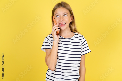 Pleasant looking happy Beautiful caucasian kid girl wearing striped  T-shirt has nice telephone conversation and looks aside, has nice mood and smiles positively while talks via cell phone