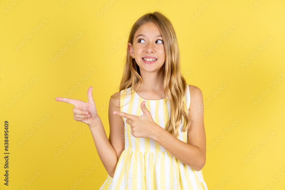 Optimistic Beautiful caucasian kid girl wearing yellow dress points with both hands and looking at empty space. Optimistic Beautiful caucasian kid girl wearing yellow dress points with both hands and looking at empty space.