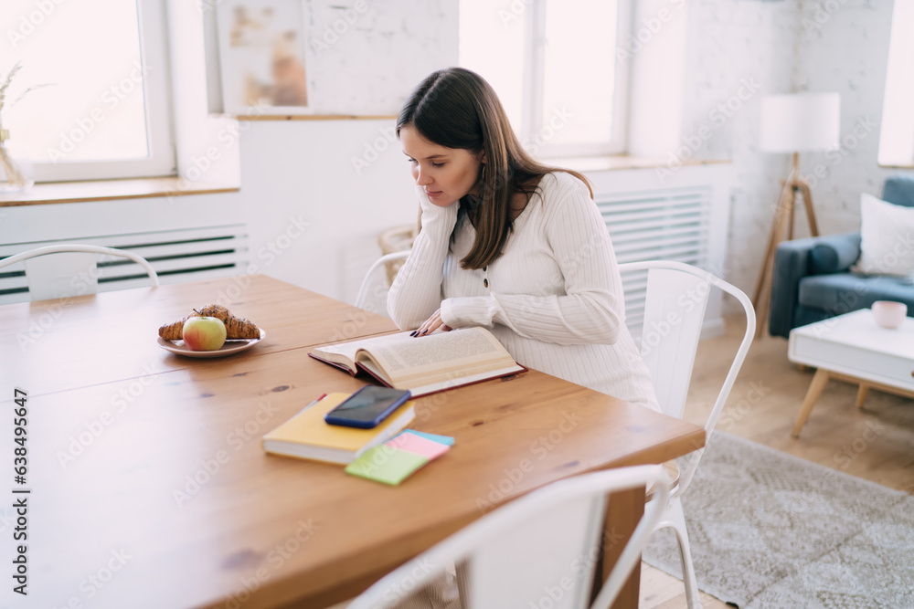 Focused pregnant woman sitting at table and reading book in kitchen Focused pregnant woman sitting at table and reading book in kitchen