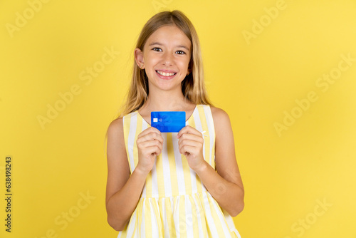 Photo of cheerful Beautiful caucasian kid girl wearing yellow dress hold debit card look empty space