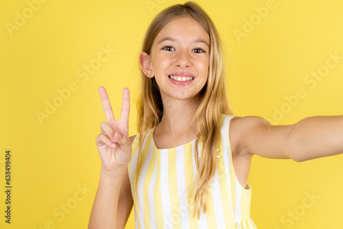 Positive Beautiful caucasian kid girl wearing yellow dress take selfie make v-sign