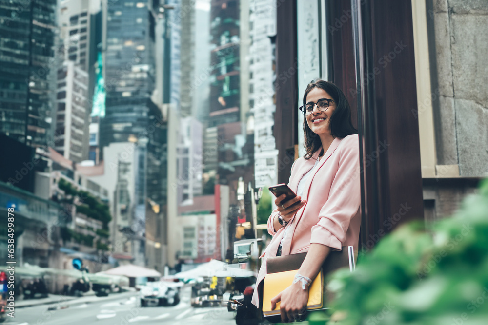 Cheerful busy woman with smartphone and papers standing next to building Cheerful busy woman with smartphone and papers standing next to building
