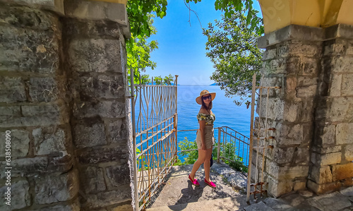 A woman in a straw entering the beach in Lovran, Croatia. The Mediterranean Sea is calm. There is a stony arch above her and a metal gate. She is smiling, enjoying the summer holidays. Happiness photo