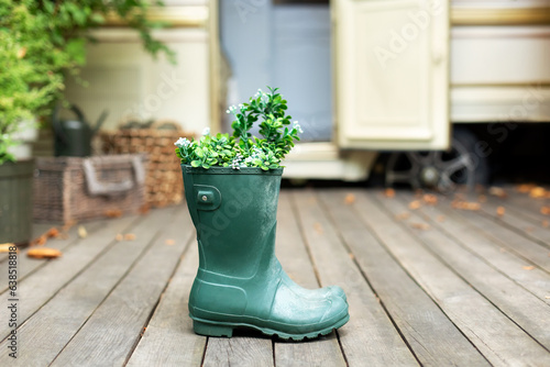 Green rubber boots with flowers and plants bouquet to contryyard of house. Cozy decor of autumn terrace of house. Garden boots. Rustic composition decor. Rain boots in front of house on porch. 