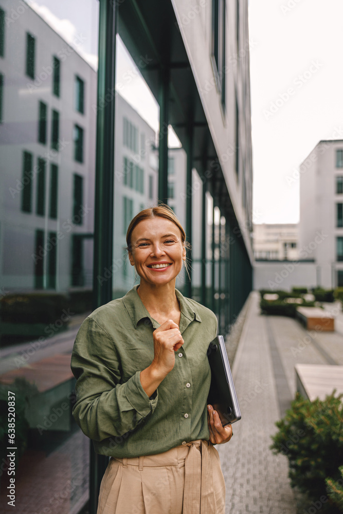 Fototapeta premium Businesswoman with laptop is standing on modern office building background and looking at camera