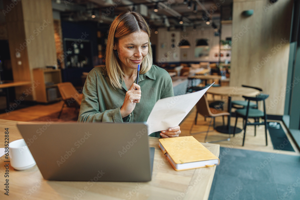 Focused woman analyst working with documents and use laptop while sitting in cozy coworking Focused woman analyst working with documents and use laptop while sitting in cozy coworking