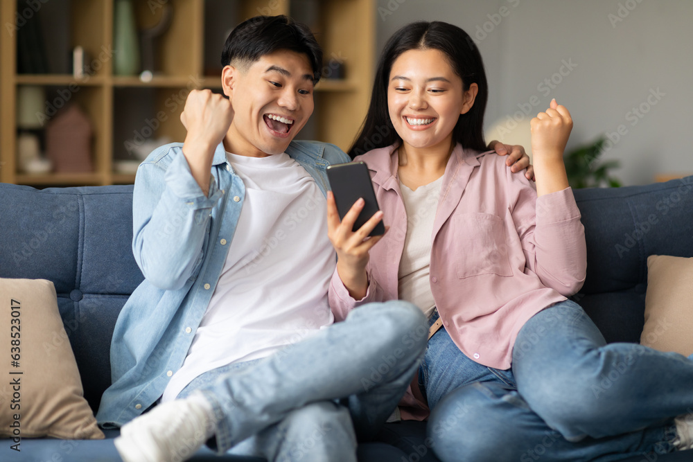 Joyful Asian Couple Celebrating Success Looking At Smartphone At Home Joyful Asian Couple Celebrating Success Looking At Smartphone At Home