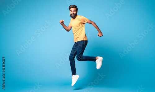 Excited middle aged indian man running posing in mid air jumping over blue background  full length  free space