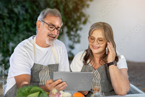 Glad old caucasian husband and wife in aprons use tablet, making phone calls at table in garden at home