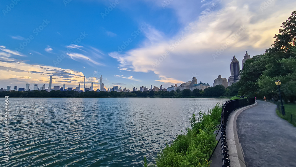 Fototapeta premium Modern cityscape of New York framed with trees with striking skyscrapers and greenery against a vibrant sky, seen from Jacqueline Kennedy Oasis Reservoir. There is a walkway along the lake. Calm