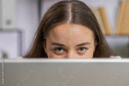 Confident business woman hiding behind laptop computer, looking at camera, spying his colleagues working, peeping. Professional freelancer girl looking from behind computer with cunning eyes glance