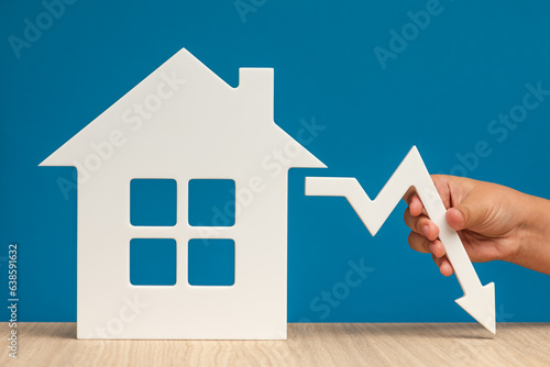 The collapse in real estate prices. Sale in the housing market. Reducing the value of real estate. Model of a house on a blue background and hand holding a white graph arrow pointing down.