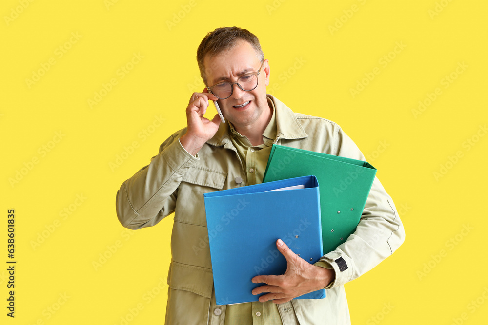 Stressed mature man with folders talking by mobile phone on yellow background. Deadline concept Stressed mature man with folders talking by mobile phone on yellow background. Deadline concept