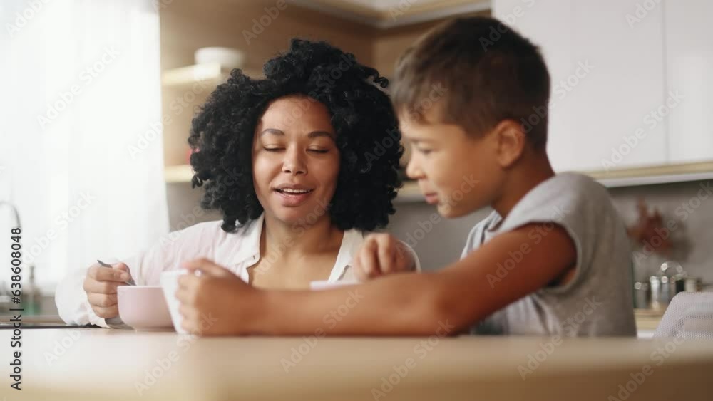 Smiling young mother with cute little son kid enjoying beautiful morning with tasty breakfast hold smartphone watching online videos entertainment program in social media together in kitchen at home Smiling young mother with cute little son kid enjoying beautiful morning with tasty breakfast hold smartphone watching online videos entertainment program in social media together in kitchen at home