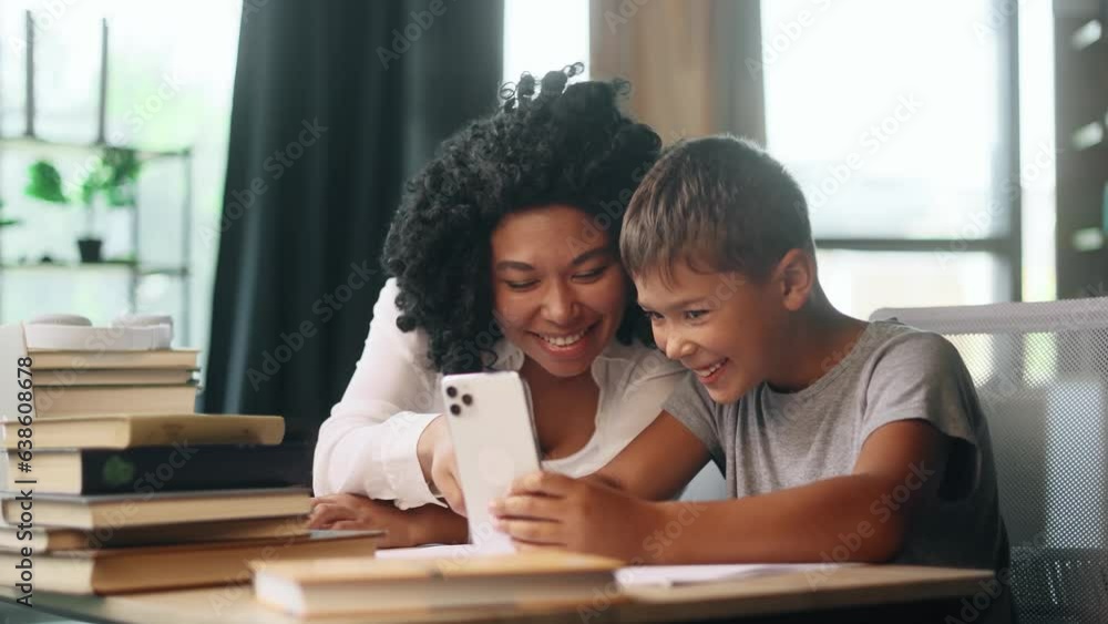 Smiling young mother with cute little son kid take break of education and hold smartphone watching online funny videos entertainment program in social media together at desk at home Smiling young mother with cute little son kid take break of education and hold smartphone watching online funny videos entertainment program in social media together at desk at home