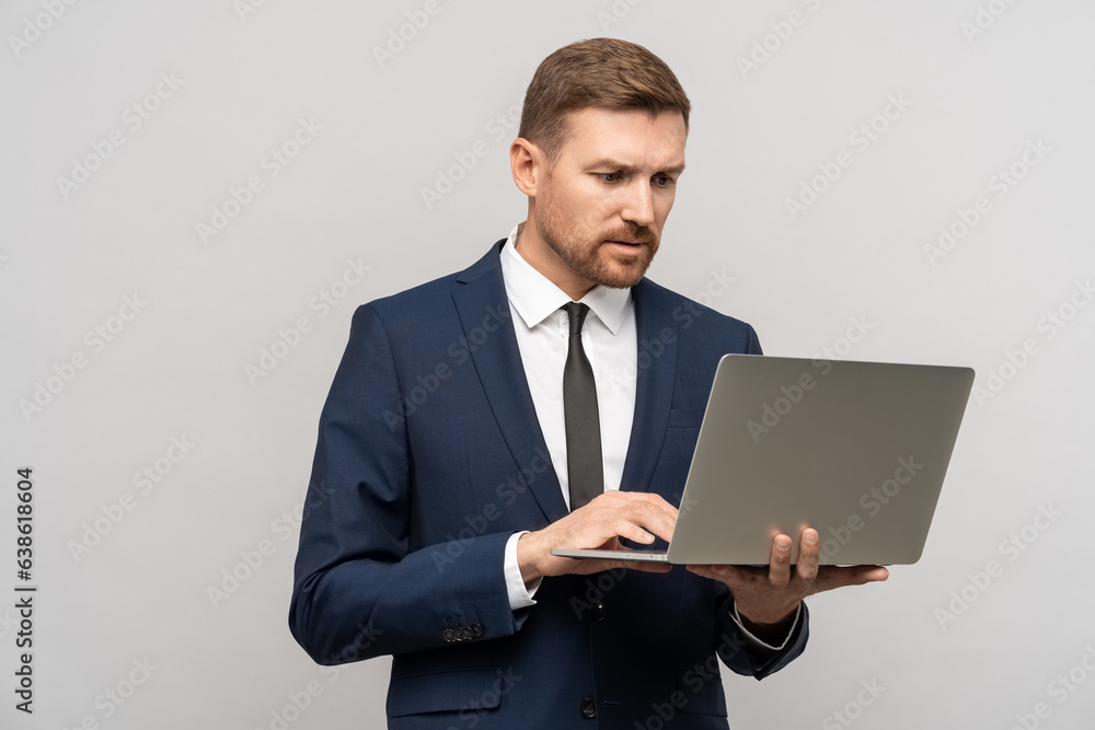 Serious frowning businessman in suit looking at laptop screen on grey background. Office work, job. Problems, troubles at work, mistakes, errors in documents, bad news, negative reactions concept. Serious frowning businessman in suit looking at laptop screen on grey background. Office work, job. Problems, troubles at work, mistakes, errors in documents, bad news, negative reactions concept.