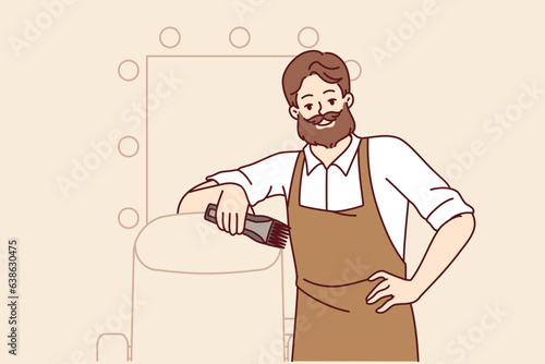 Bearded man hairdresser works in barbershop and smiles, standing near chair for client and mirror. Successful stylist in apron posing indoors at barbershop and holding trimmer in hand
