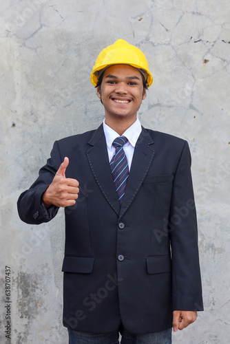 Happy Asian businessman in suit wearing safety yellow helmet smiling and give thumb up to the camera