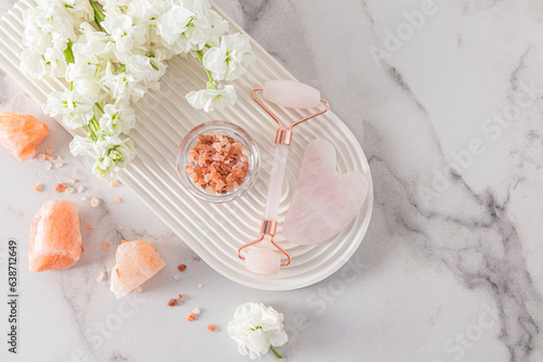 A rose quartz cosmetic roller massager, a gua sha scraper and flowers lie on a white podium in the form of an arch. Top view. self-care. photo