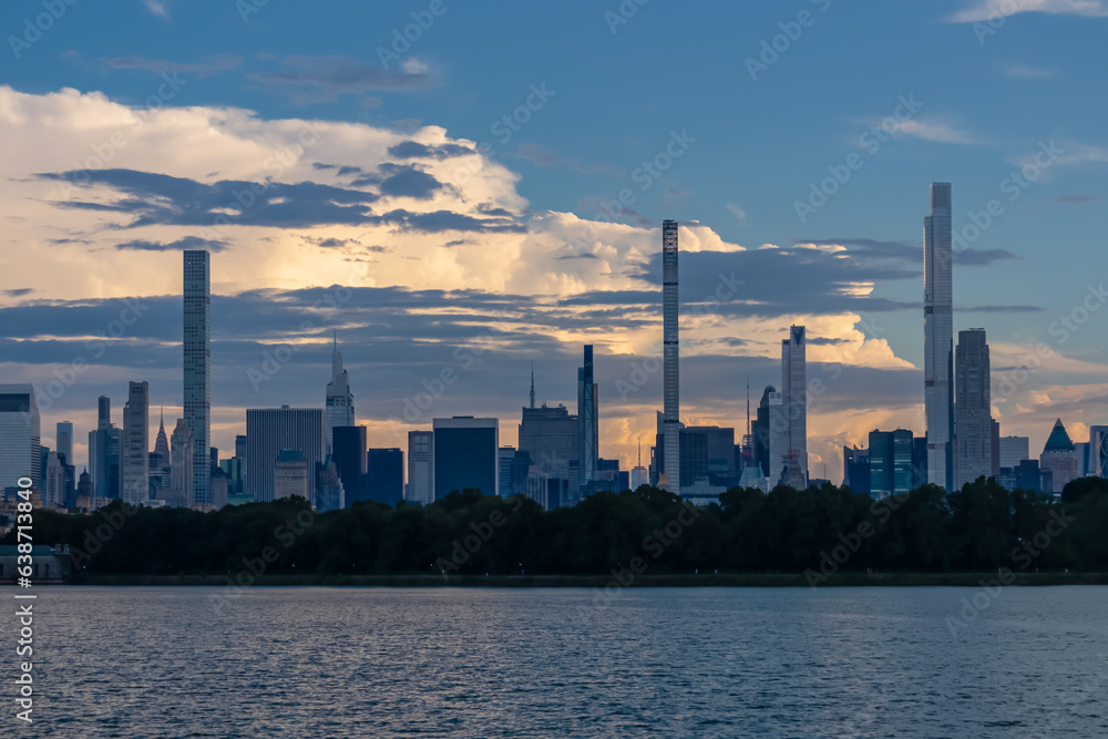 Captivating New York urban skyline at dusk with striking and modern skyscrapers reflecting on water seen from Jacqueline Kennedy Oasis Reservoir. The buildings are taller than surrounding clouds.