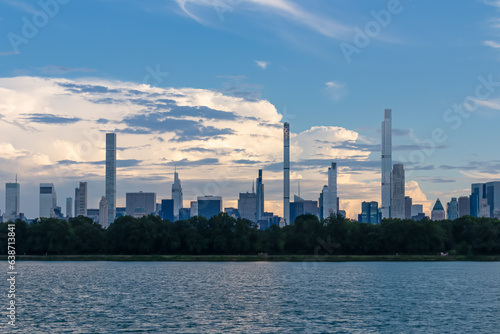 Captivating New York urban skyline at dusk with striking and modern skyscrapers reflecting on water seen from Jacqueline Kennedy Oasis Reservoir. The buildings are taller than surrounding clouds.