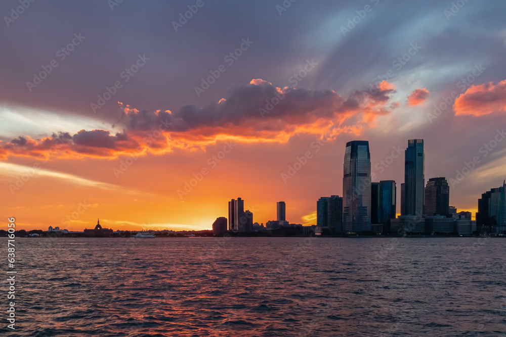 Breath-taking afterglow over New York City skyline and Hudson River at dusk. The sun goes down behind tall skyscrapers. The sky is bursting with orange and yellow. Calmness and relaxation