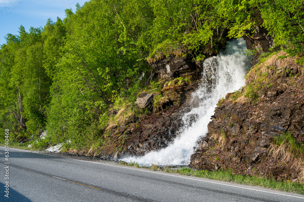 Waterfall flows from green forest just next to the asphalt road