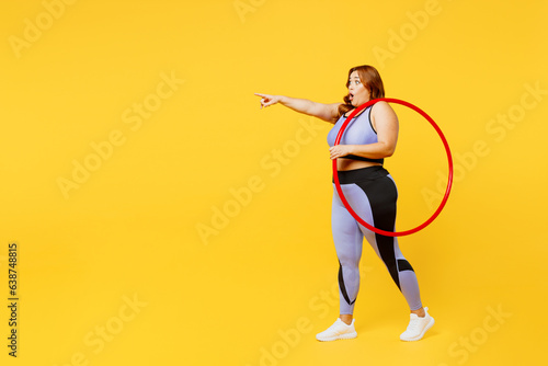 Full body side view young plus size big fat fit woman wear blue top warm up train hold hula hoop walk go point finger aside isolated on plain yellow background studio home gym. Workout sport concept.