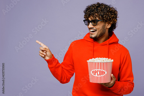 Young fun Indian man in 3d glasses wear red orange hoody casual clothes watch movie film hold bucket of popcorn in cinema point aside isolated on plain pastel purple color background studio portrait.