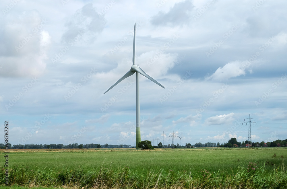 Windenergie: Ein riesiges Windrad in einer ländlichen Gegend mit bewölktem Himmel und mehreren Strommasten nebst Leitung im Hintergrund, viel Copy Space Windenergie: Ein riesiges Windrad in einer ländlichen Gegend mit bewölktem Himmel und mehreren Strommasten nebst Leitung im Hintergrund, viel Copy Space