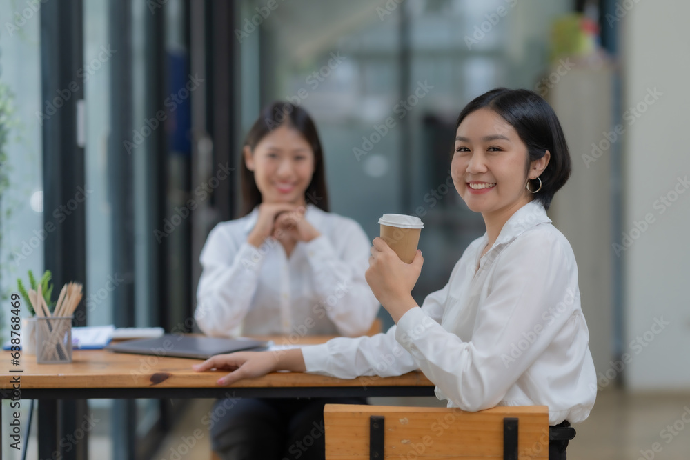 Side view of Asian woman holding coffee cup looking at camera with colleague working in background at office.