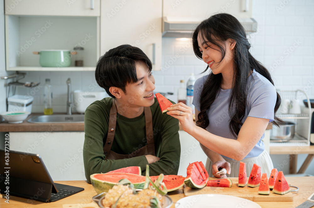A happy Asian man is being fed a slice of watermelon by his girlfriend in the kitchen