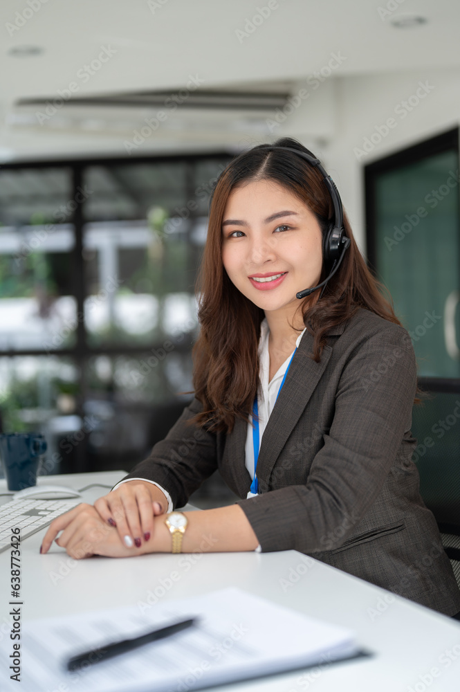 A charming Asian female sales agent with a headset sits at her desk and looks at the camera