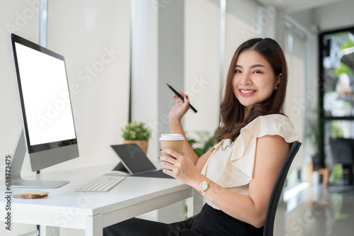 A gorgeous Asian businesswoman sits in front of a computer with a coffee cup in her hand