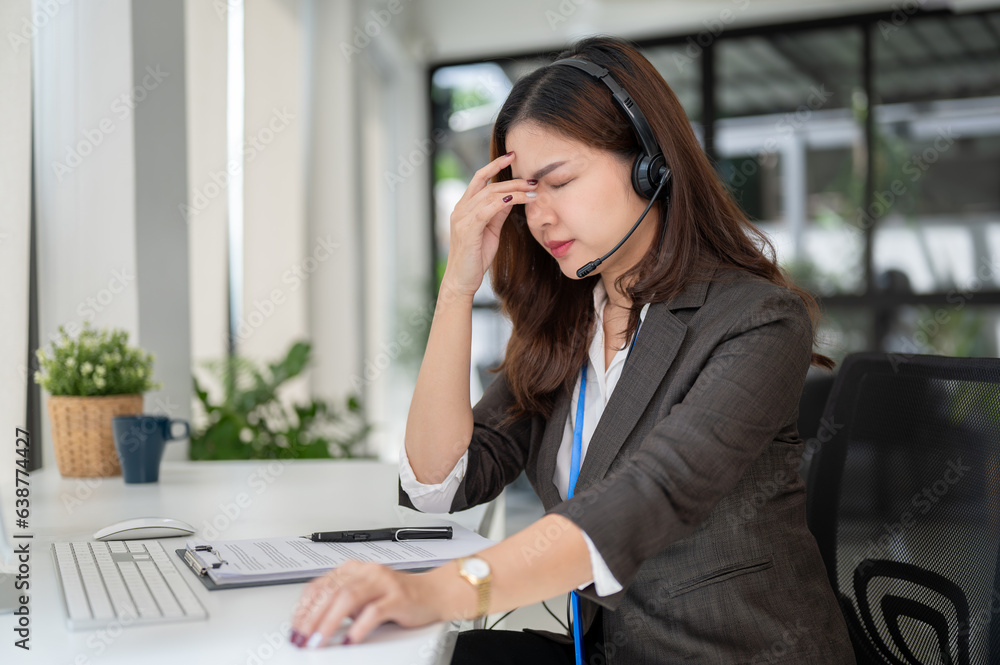 A stressed and tired Asian female centre operator sits at her desk, suffering from headaches A stressed and tired Asian female centre operator sits at her desk, suffering from headaches
