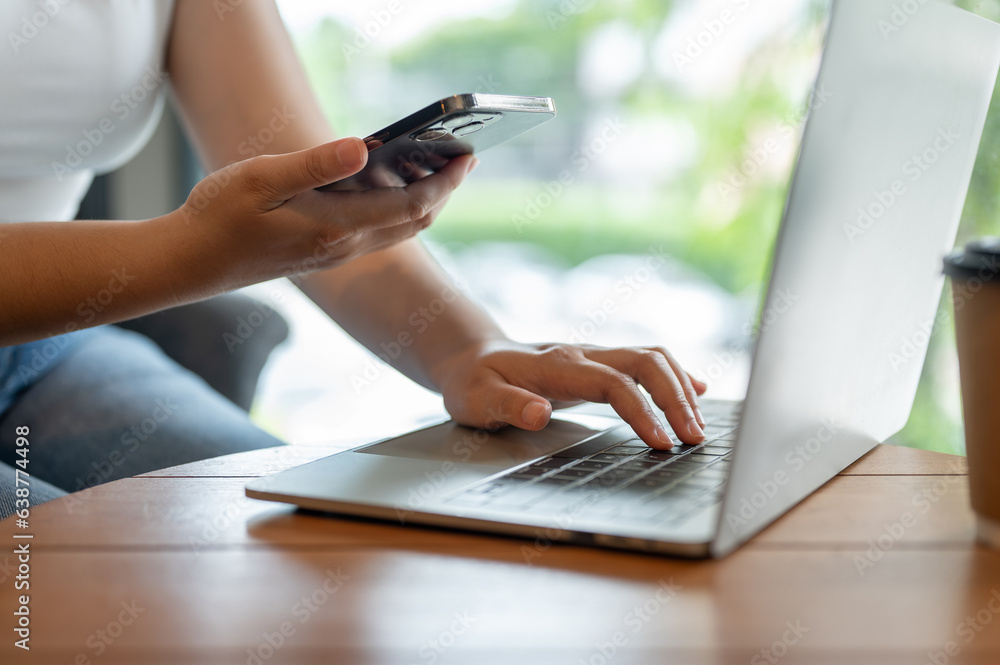 Fototapeta premium A female freelancer working remotely at a cafe, using her smartphone and laptop at a table.