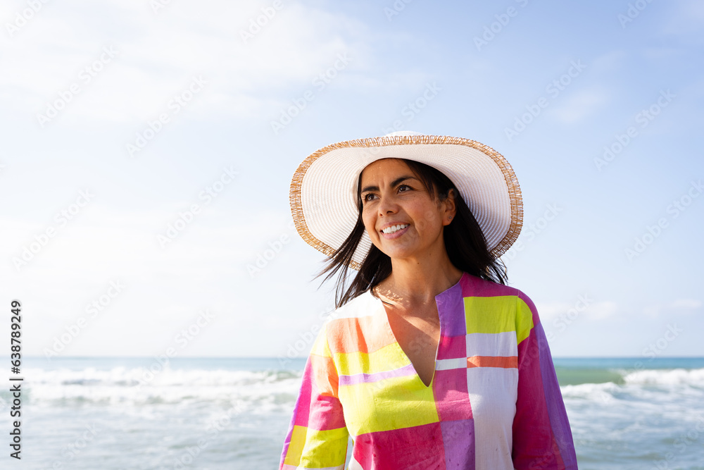 Gorgeous latina woman in a hat and colorful dress walking along the beach on a lovely bright and radiant spring day