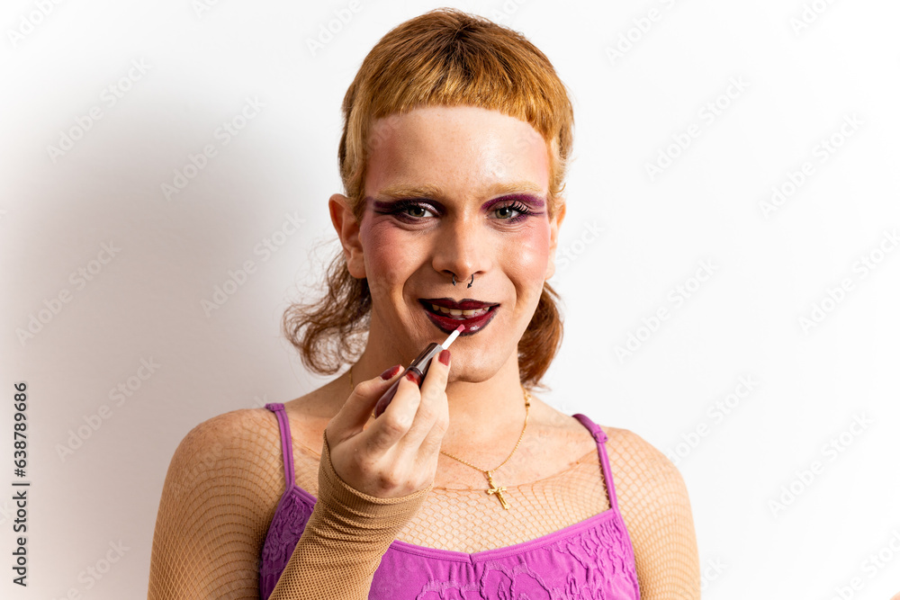 Portrait of a red-haired non-binary person is posing on a white background while painting his lips and looking at the camera happily. Concept of people is not identified with any gender. Men in makeup Portrait of a red-haired non-binary person is posing on a white background while painting his lips and looking at the camera happily. Concept of people is not identified with any gender. Men in makeup