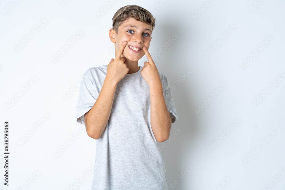 Happy Beautiful kid boy wearing grey casual t-shirt with toothy smile, keeps index fingers near mouth, fingers pointing and forcing cheerful smile Happy Beautiful kid boy wearing grey casual t-shirt with toothy smile, keeps index fingers near mouth, fingers pointing and forcing cheerful smile