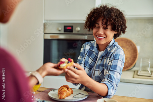 A happy teenage boy sharing a piece of bread with his mother at the dining table.