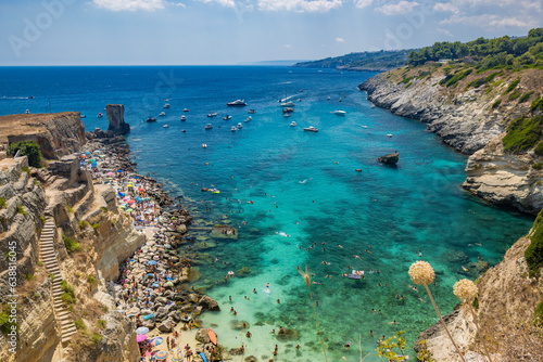 The amazing bay of Porto Miggiano, in Santa Cesarea Terme, resort of Salento, Puglia, Italy. Full of tourists and boats, in the clear, blue and turquoise sea. Small beach enclosed between high cliffs