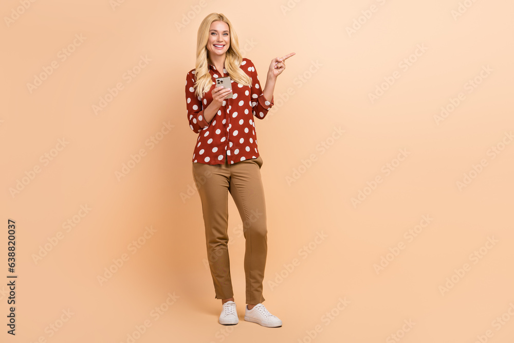 Full size photo of charming nice girl hold smart phone indicate finger empty space isolated on beige color background Full size photo of charming nice girl hold smart phone indicate finger empty space isolated on beige color background