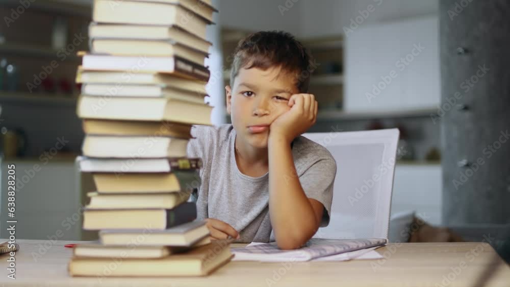 Tired exhausted stressed little boy student looking from behind large pile of books and looking at camera with sad face while preparing for exams at home Overwork education concept Tired exhausted stressed little boy student looking from behind large pile of books and looking at camera with sad face while preparing for exams at home Overwork education concept