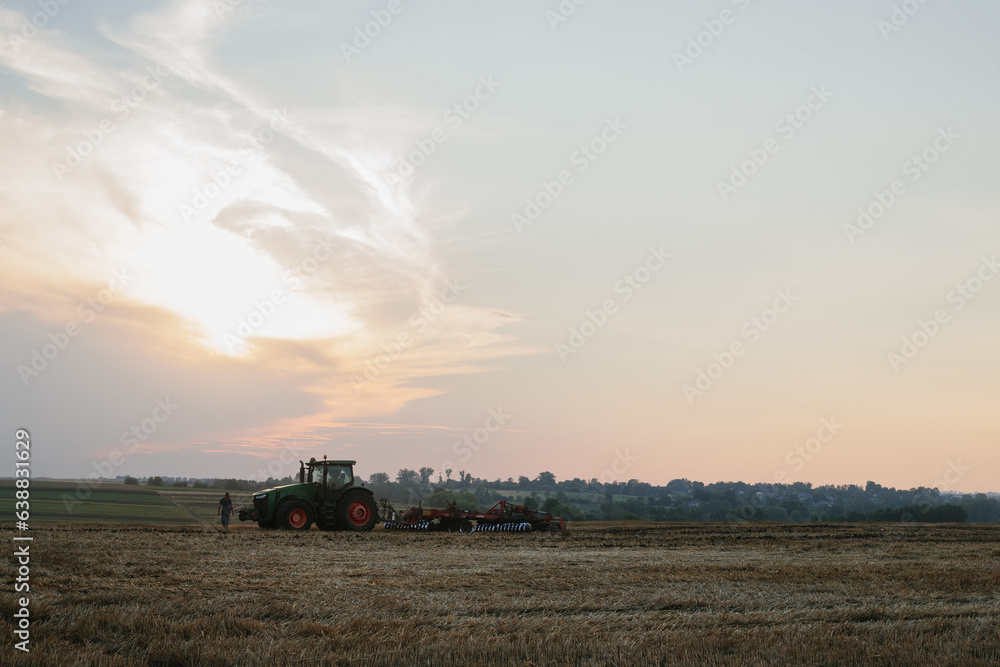 Fototapeta premium A modern tractor with a heavy trailed disc harrow plows a wheat field at sunset.