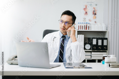 Asian Doctor talking over his mobile phone and making notes in a note. Medicine professional talking on phone and clinical report test results.