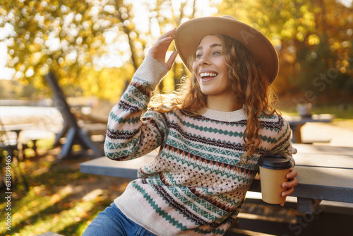 Portrait of a cute woman in a hat sits with a cup of coffee overlooking the lake. Outdoor photo of relaxed curly woman enjoying hot drink in autumn park.