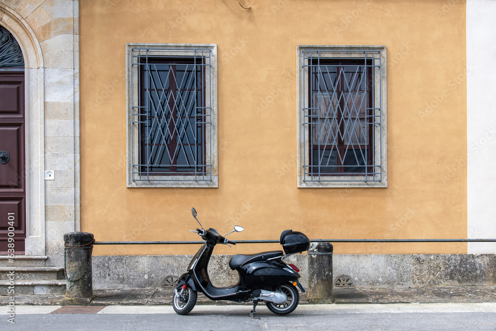 Black scooter parked on sidewalk in front of a historical Italian house with yellow plastered walls, stone pillars and large wooden door