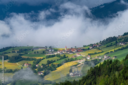 Panoramic view on village of Aicha or Aica in South Tyrol, Italy with characteristic church steeple surrounded by pasture and low hanging white clouds surrounding mountains of the Dolomites photo