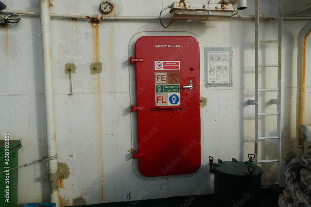 Red metal door with stickers is closing the entrance to safety locker on the container vessel. The wall in aft station of the ship are partly rusted has white colour.
Red metal door with stickers is closing the entrance to safety locker on the container vessel. The wall in aft station of the ship are partly rusted has white colour.